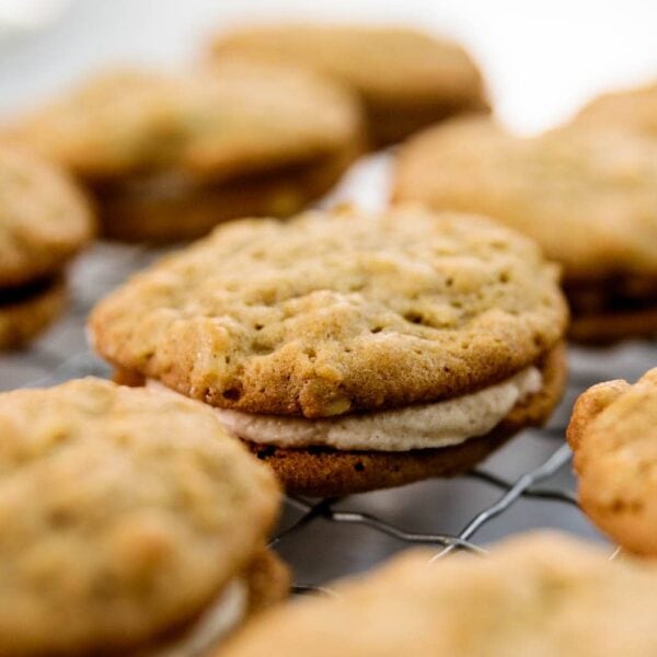 Two oatmeal spice cookies sandwiching a layer of white creme filling with multiple cookies in background all on a wire cooling rack on a marble surface