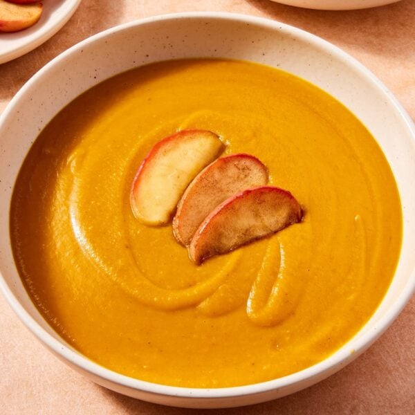 Large white bowl filled with orange colored squash soup with sliced and cooked apples on top with more apples in background.