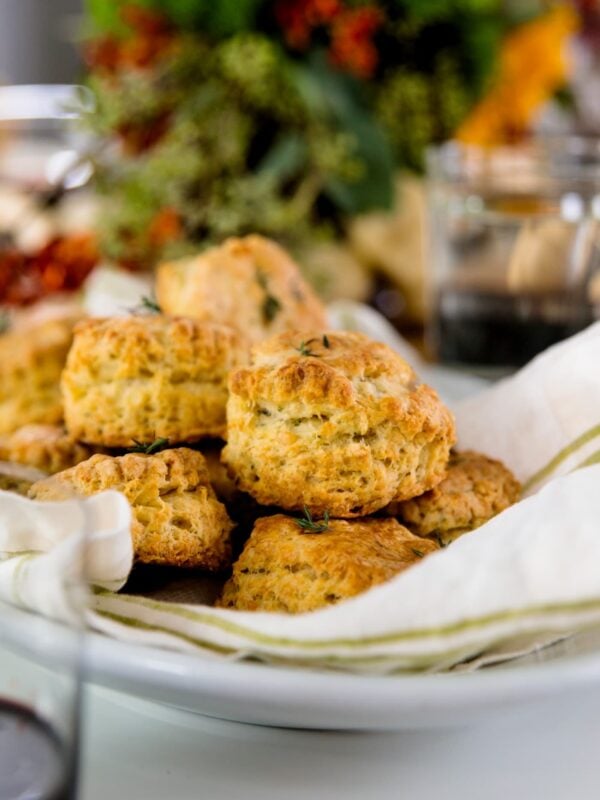 Herb cheese biscuits sitting on white and green linen in white bowl with flowers in background and glass in foreground all on white surface
