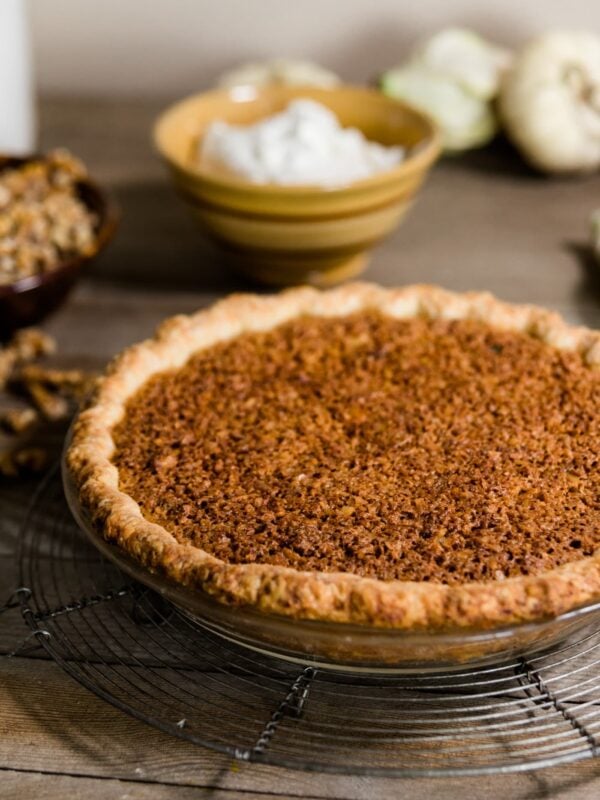 Oatmeal nut pie sitting on wire cooling rack with whipped cream, white pumpkins, sugar and toasted walnuts in background on wood plank surface
