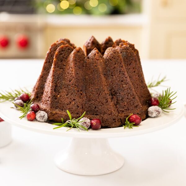 Tall brown cake sitting on white cake plate with cranberries sprinkled around all on white surface with oven and cabinetry in background