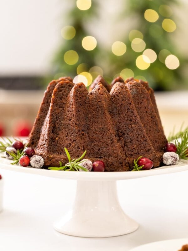 Tall brown cake sitting on white cake plate with cranberries sprinkled around all on white surface with oven and cabinetry in background