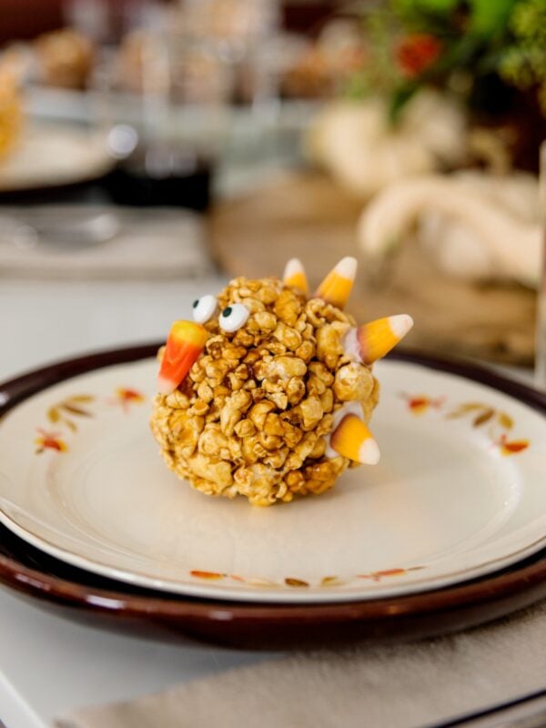 Turkey-shaped popcorn balls sitting on white and maroon plates on white Thanksgiving table set with pumpkins