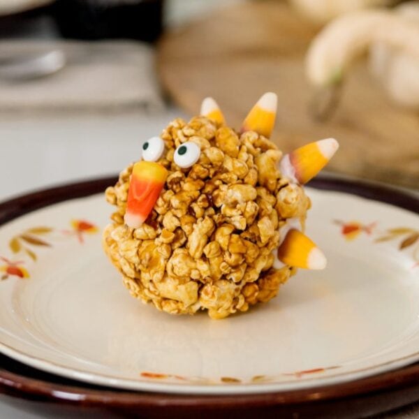 Turkey-shaped popcorn balls sitting on white and maroon plates on white Thanksgiving table set with pumpkins