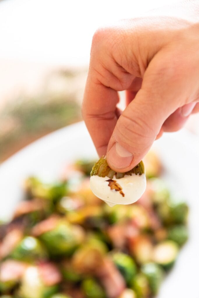 Hand holding green Brussels sprout with white creamy sauce on the end with rest of Brussels sprouts in background