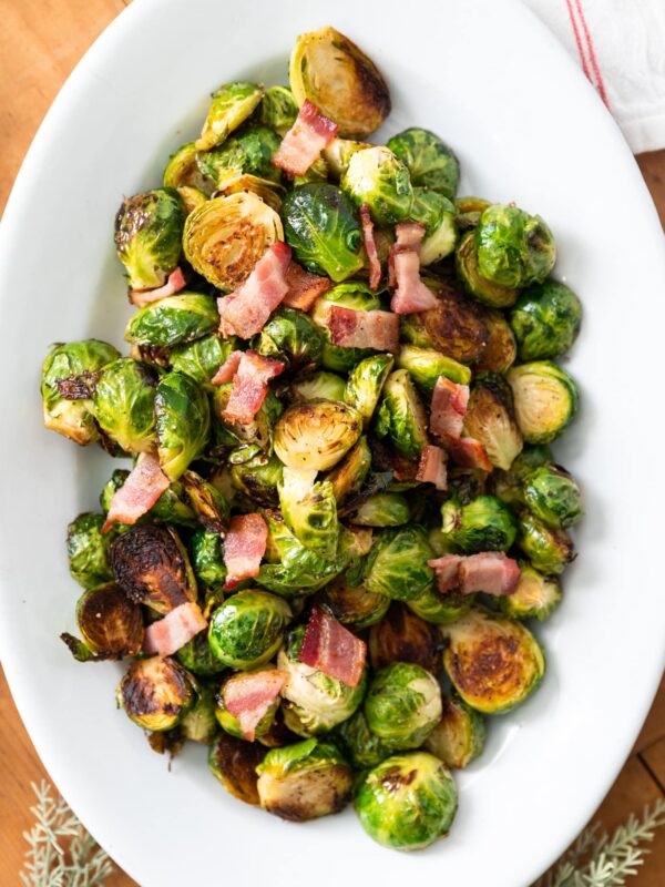Top down view of Brussels sprouts in white bowl topped with bacon sitting on wood board