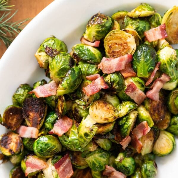 Top down view of green Brussels sprouts sitting in white platter topped with bacon sitting on wood board with greenery