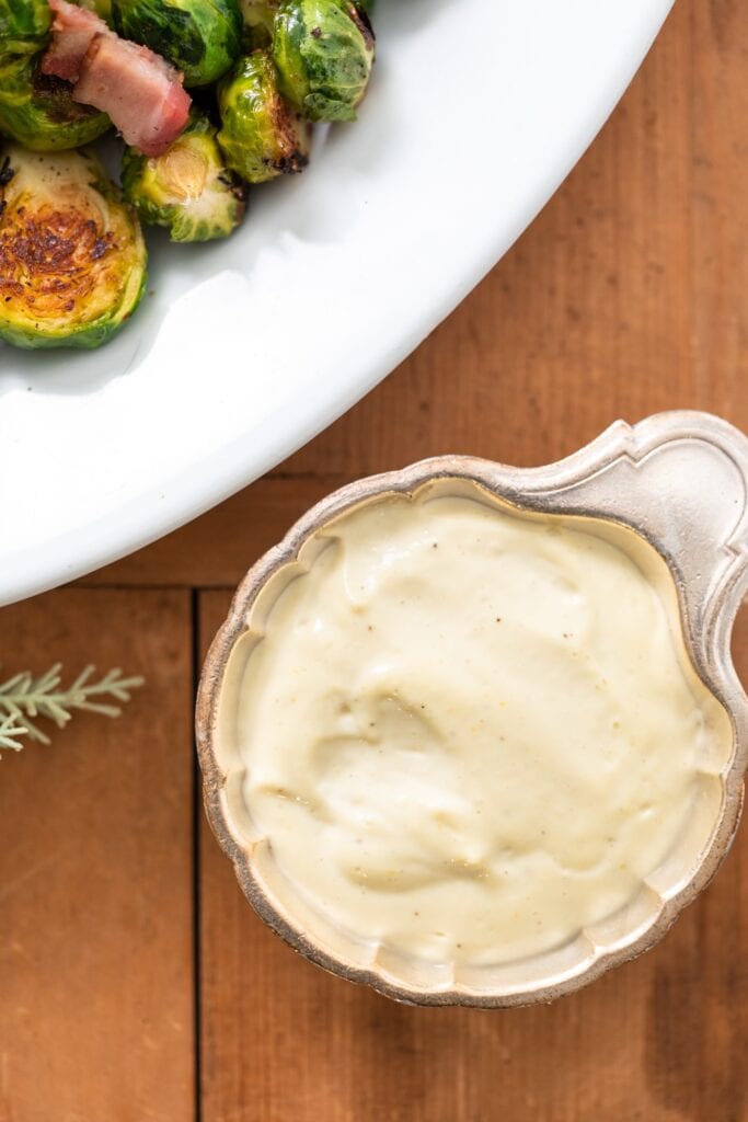 Silver container filled with white colored garlic aioli sitting on wood surface with white bowl filled with Brussels sprouts and bacon
