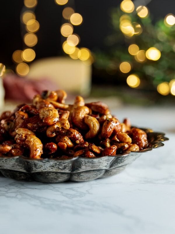 Silver tray filled with candied curry nuts all on white marble surface with twinkling Christmas lights in the background.