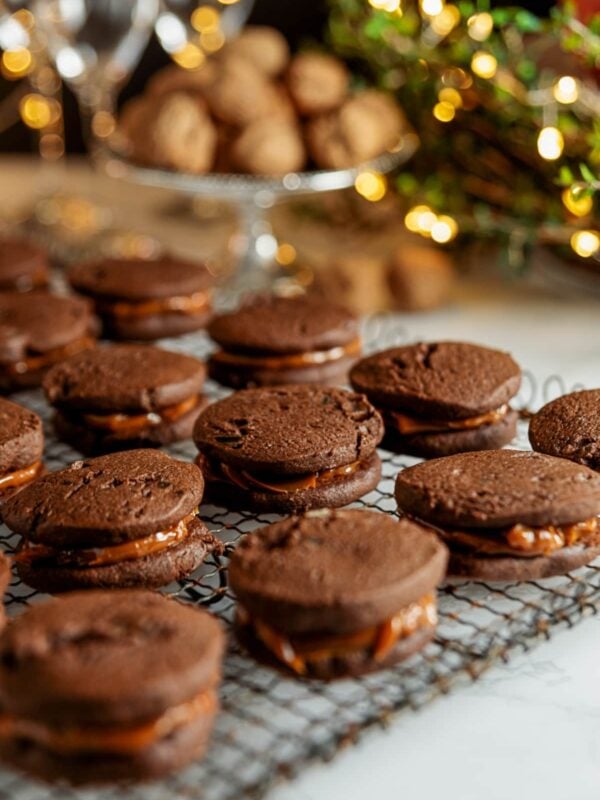 Chocolate, ginger, and dulce de leche sandwich cookies sitting on a wire cooling rack on a white marble surface with Christmas lights in the background.