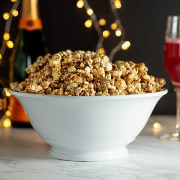 Cinnamon and cayenne caramel corn in white bowl with bottle of champagne, pink drink in flute and twinkling lights in background on marble surface