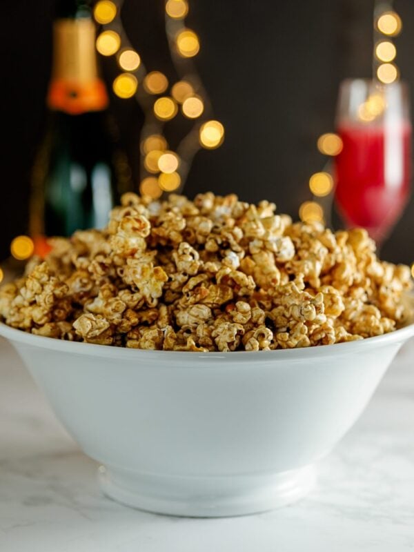 Cinnamon and cayenne caramel corn in white bowl with bottle of champagne, pink drink in flute and twinkling lights in background on marble surface