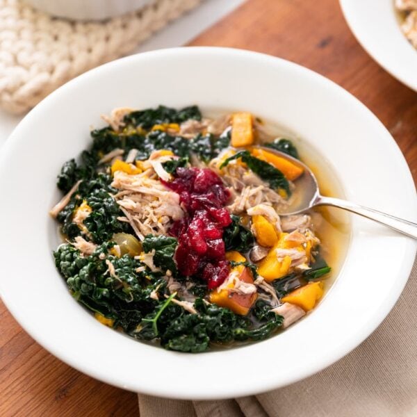 White bowl filled with turkey and kale soup with spoon sitting in broth all on wood surface with extra bowl of soup to the side