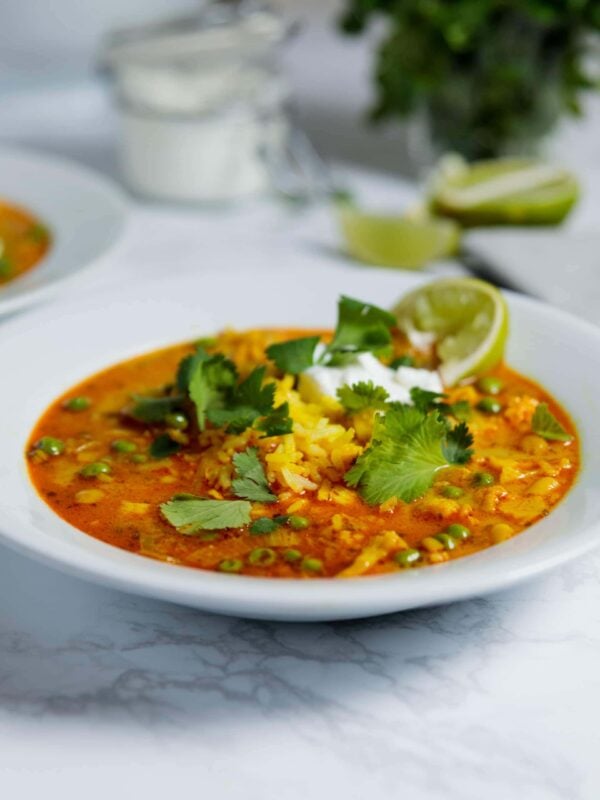 White bowl filled with orange colored cauliflower curry soup with cilantro leaves on top as well as slice of lime all on white surface