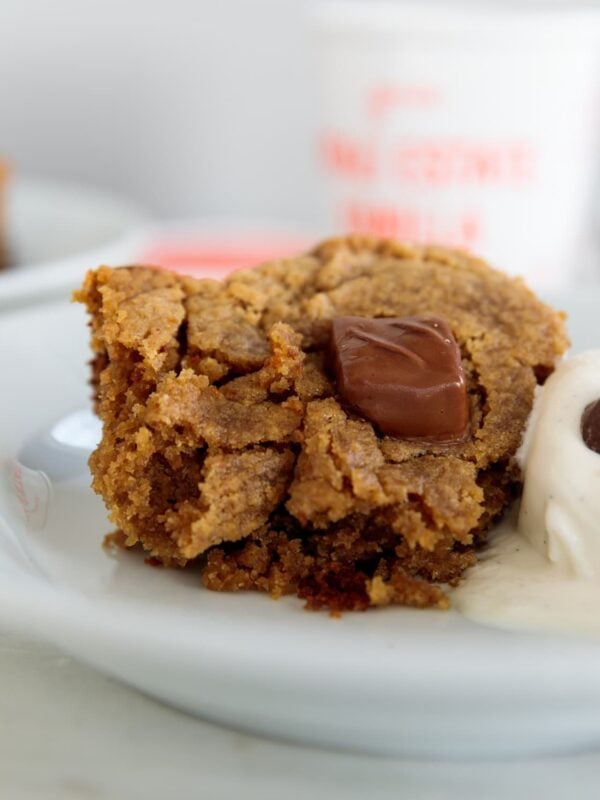 Piece of peanut butter cake with chocolate candies on top served beside a scoop of vanilla ice cream on a plate.