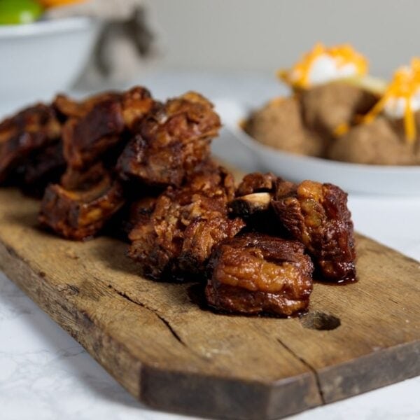 Wooden cutting board on white marble surface with short ribs with homemade bbq sauce and baked potatoes in background.