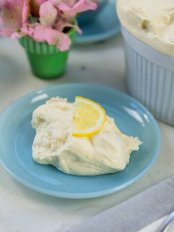 Blue plate with serving of lemon pudding soufflé with slice of lemon on top all on white marble surface with pink flowers in the background.