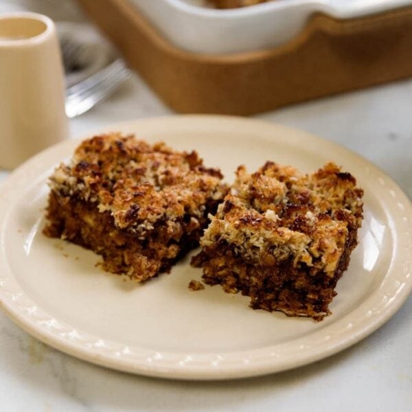 Oatmeal cake with two pieces sitting on a tan plate with baking dish with more cake in background.