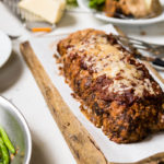 Pizza meatloaf on piece of parchment paper on a cutting board with green beans to the side.