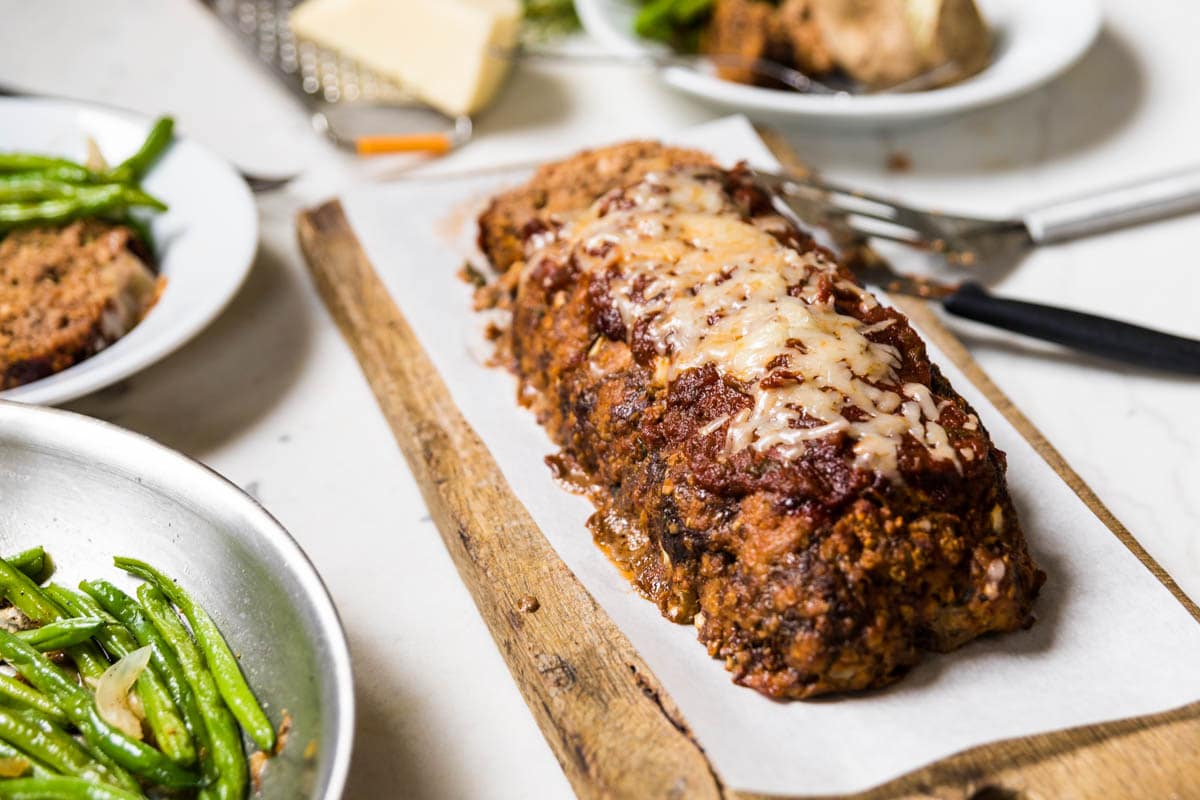 Pizza meatloaf on piece of parchment paper on a cutting board with green beans to the side.