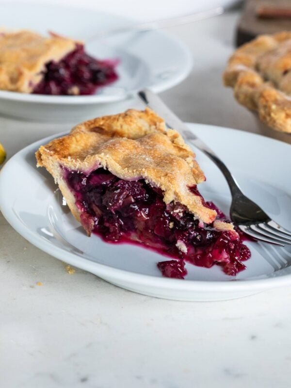 White plate with slice of rhubarb blueberry pie with fork resting on plate with rest of pie in the background.