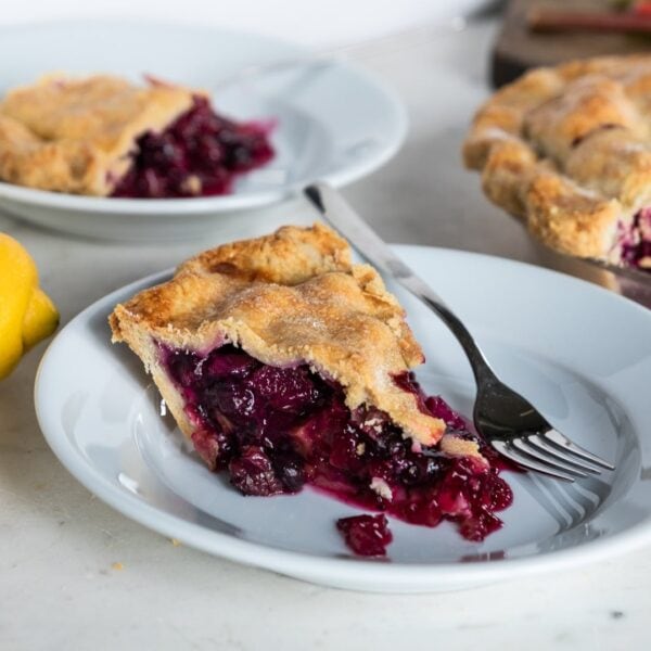 Rhubarb and blueberry pie on a white plate with fork with second plate of pie and pie dish in background with lemons on a marble surface