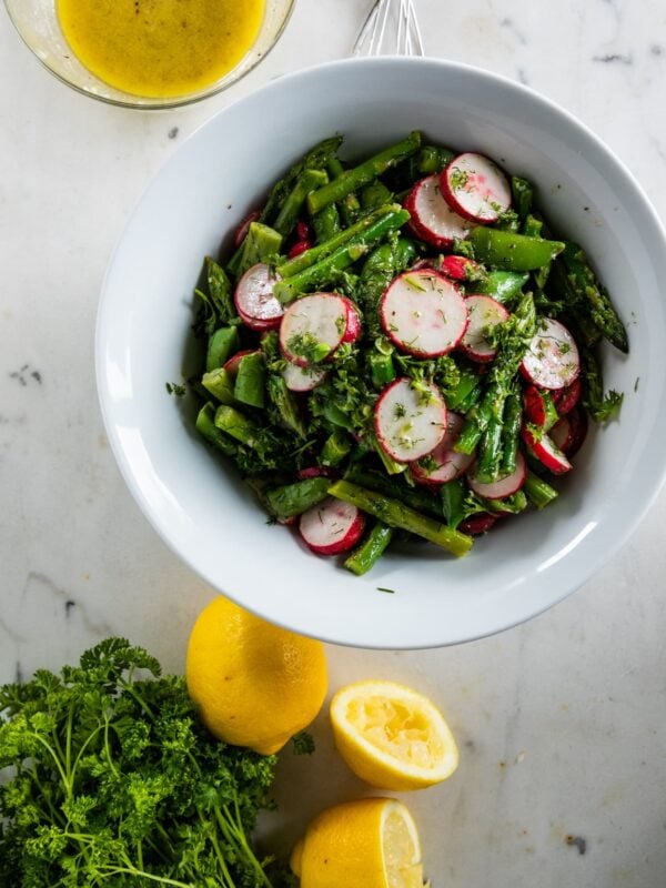 Top down view of asparagus salad with radish and sugar peas in a large white bowl with extra lemons, parsley, garlic and homemade vinaigrette on a marble surface