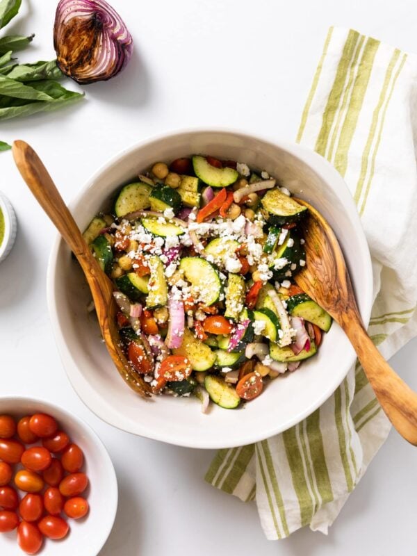 Top down view of white bowl holding zucchini and tomato salad all sitting on white countertop with grilled onion and extra tomatoes