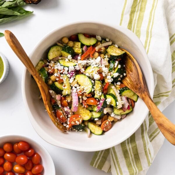 Top down view of white bowl filled with zucchini salad with wooden tongs all on white countertop with extra basil dressing and tomatoes