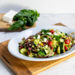 Grilled zucchini and onion salad in large white bowl on top of wood cutting board with salt and pepper, feta and basil in background all on a marble surface