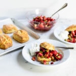 Strawberry and rhubarb shortcakes with whipped cream on two bowls with golden biscuits and macerated fruit in a bowl in the background on a marble surface
