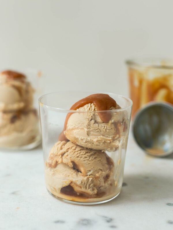 Caramel macchiato ice cream with caramel sauce drizzled on top in a clear glass with a second glass full of ice cream in background as well as extra homemade caramel sauce in a jar and an ice cream scoop on a marble surface
