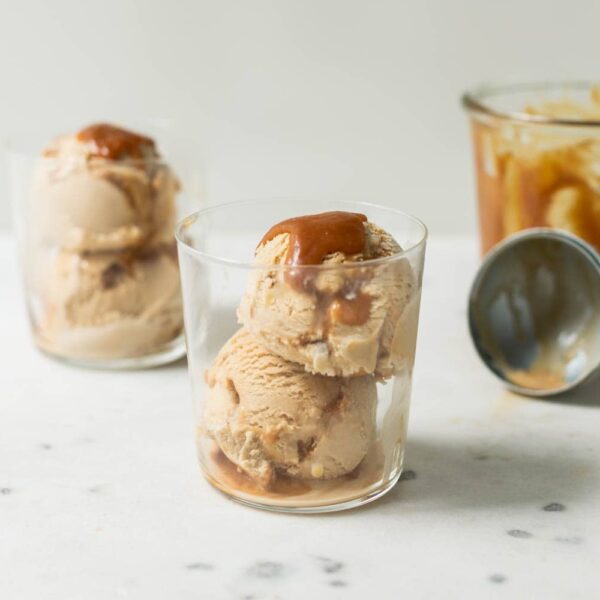 Caramel macchiato ice cream with caramel sauce drizzled on top in a clear glass with a second glass full of ice cream in background as well as extra homemade caramel sauce in a jar and an ice cream scoop on a marble surface
