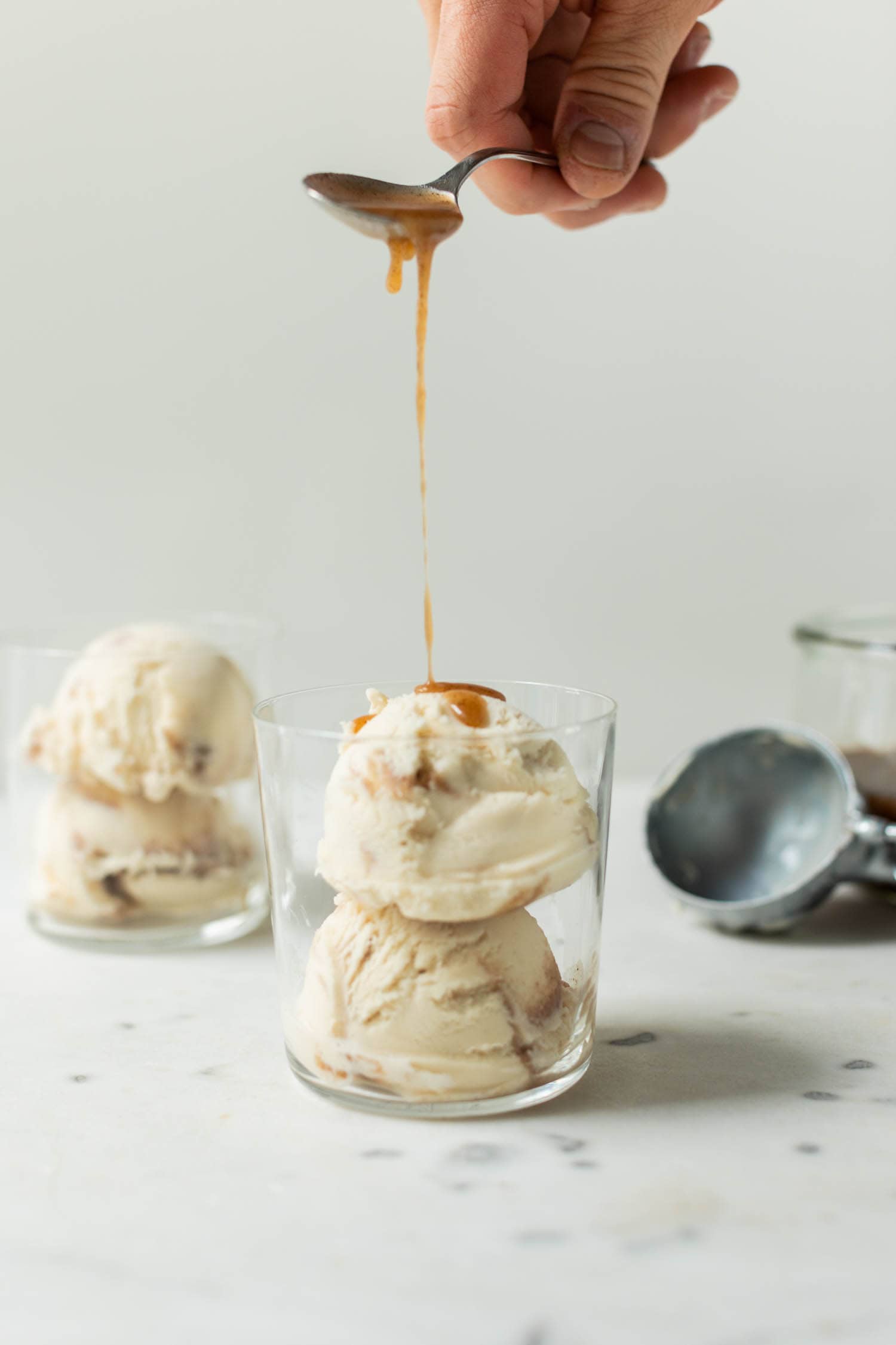 French toast ice cream drizzled with extra cinnamon sauce drizzling on top of ice cream in a clear glass on a marble surface with second ice cream cup and ice cream scoop in background