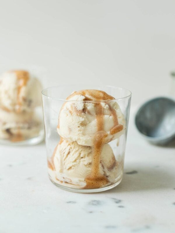 French toast ice cream drizzled with extra cinnamon sauce in a clear glass on a marble surface with second ice cream cup and ice cream scoop in background