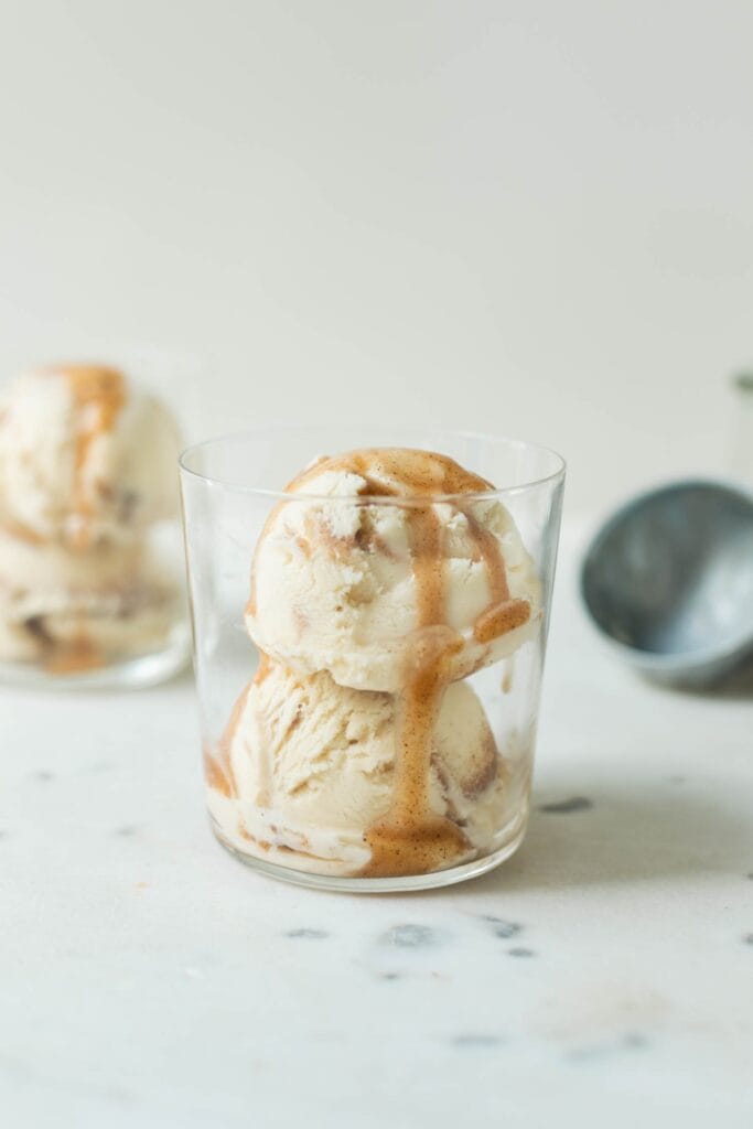 French toast ice cream drizzled with extra cinnamon sauce in a clear glass on a marble surface with second ice cream cup and ice cream scoop in background