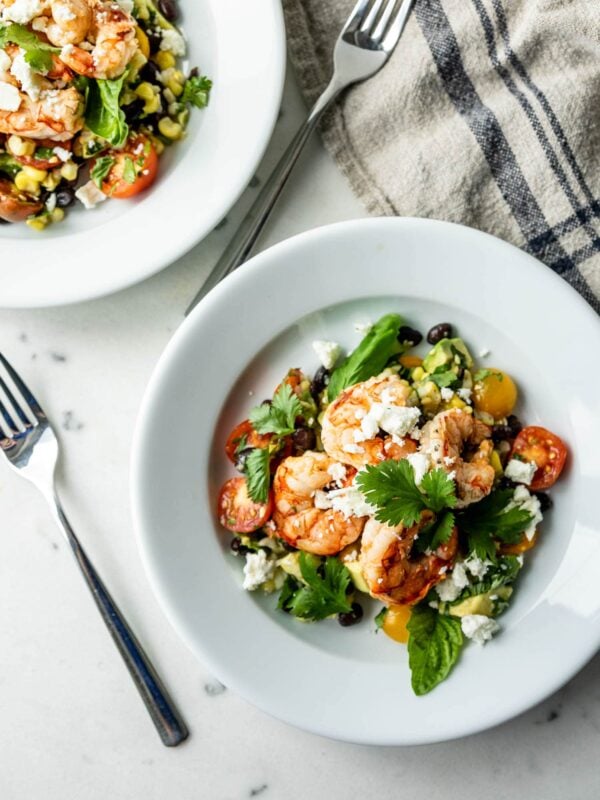 Top down view of grilled tequila lime shrimp on a bed of cherry tomatoes, corn and cilantro and sprinkled with feta cheese with a second plate, forks and linen towel in background with a marble surface