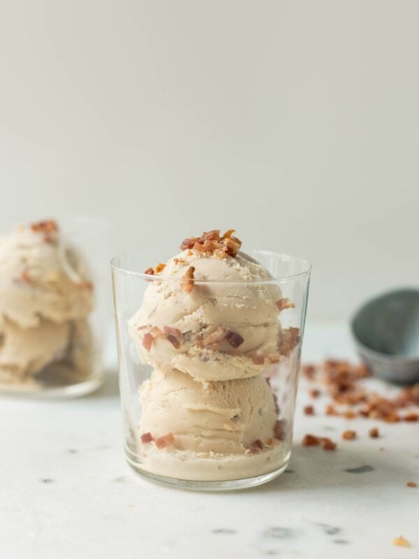 Maple bacon ice cream with bacon pieces on top and down the sides in a clear glass with a second glass, ice cream scoop and bacon pieces in background on a marble surface