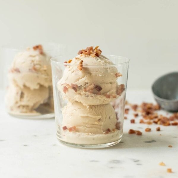 Maple bacon ice cream with bacon pieces on top and down the sides in a clear glass with a second glass, ice cream scoop and bacon pieces in background on a marble surface