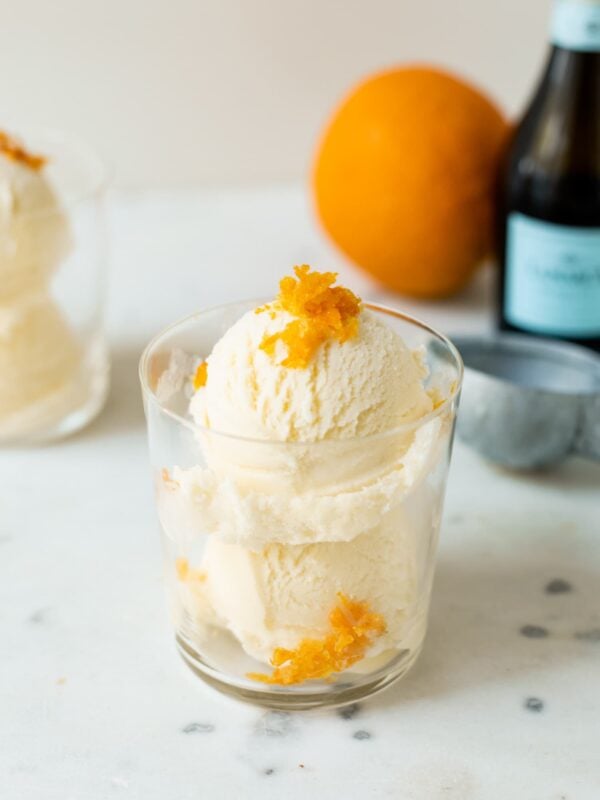 Mimosa ice cream speckled with orange zest pieces in a clear glass with second ice cream cup, orange, ice cream scoop and champagne bottle in background on a white marble surface