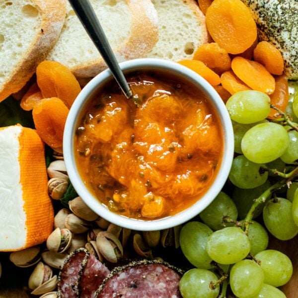 Top down view of peach jalapeño jam with a spoon in a white bowl surrounded by grapes, bread, dried apricots, cheese and goat cheese on a brown serving board