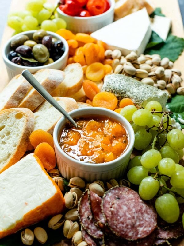 Peach jalapeño jam with a spoon in a white bowl surrounded by grapes, bread, dried apricots, cheese, olives, tomatoes and goat cheese on a brown serving board