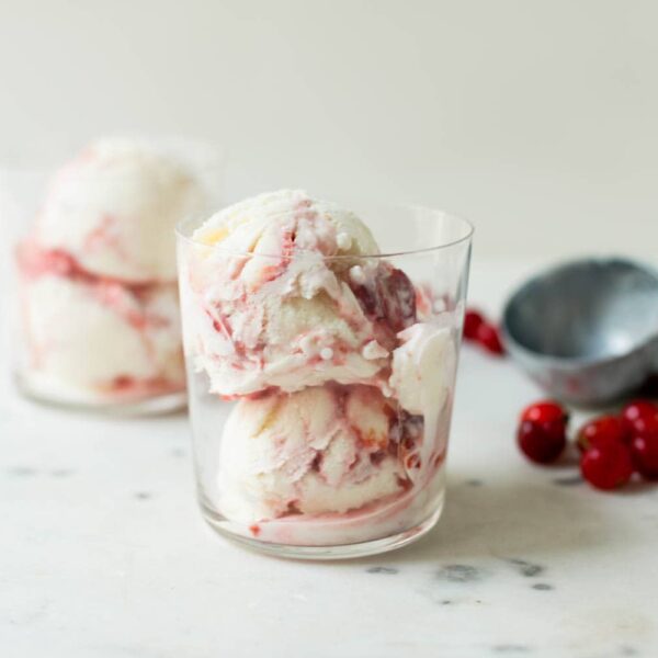 Sour cherry frozen yogurt with streaks of cherry jam throughout in clear glass with second glass, sour cherries and ice cream scoop in background on marble surface