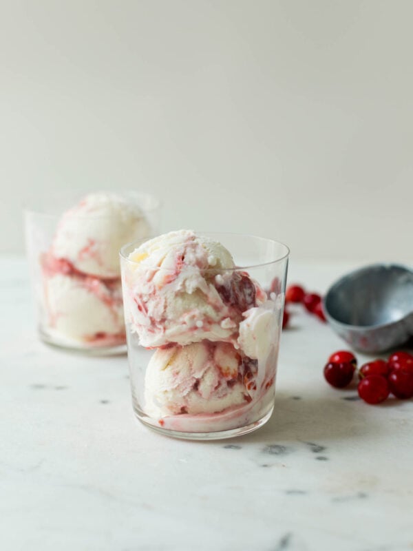 Sour cherry frozen yogurt with streaks of cherry jam throughout in clear glass with second glass, sour cherries and ice cream scoop in background on marble surface