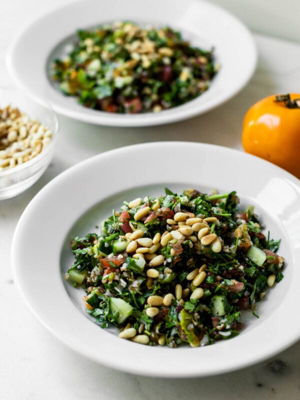 Two white bowls filled with tabbouleh topped with pine nuts with glass bowl of extra pine nuts and tomatoes to the side on a marble surface