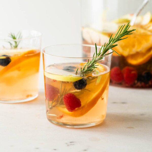 White sangria with a sprig of rosemary with blueberries, raspberries, mandarin orange pieces and apple splices with pitcher full of sangria in background on a marble surface