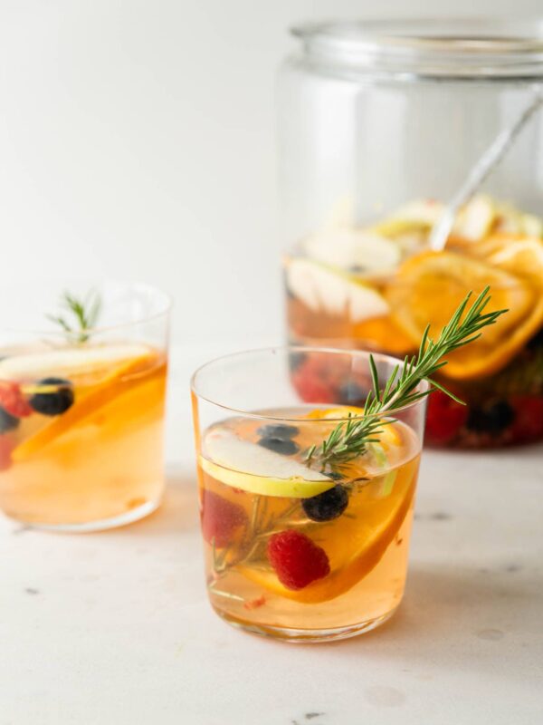 White sangria with a sprig of rosemary with blueberries, raspberries, mandarin orange pieces and apple splices with pitcher full of sangria in background on a marble surface