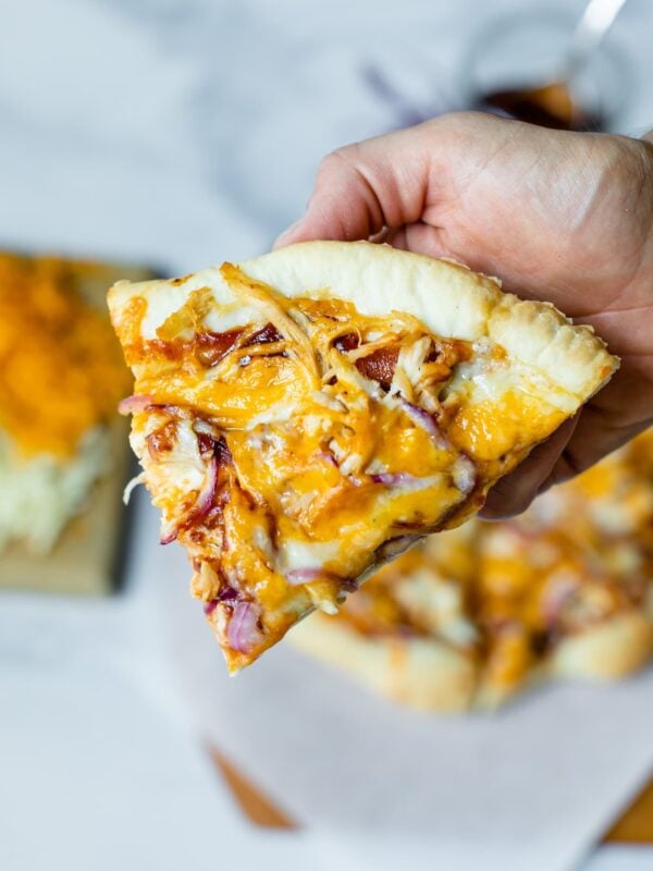 Slice of homemade barbecue chicken pizza with cheese, red onions, chicken, barbecue sauce and homemade crust being held with rest of pizza on a piece of parchment, pizza board, extra cheese on a cutting board and extra barbecue sauce in background on a marble surface