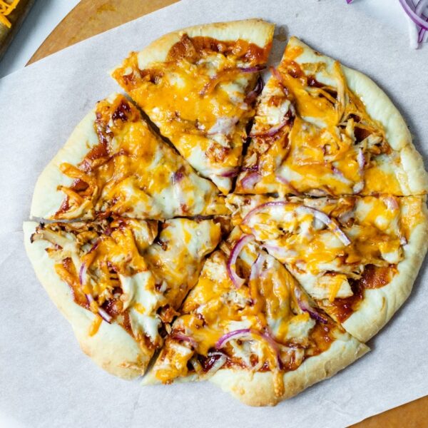 Top down view of sliced homemade barbecue chicken pizza made of chicken, cheese, barbecue sauce and red onion on a homemade crust on a piece of white parchment and pizza board with extra red onions and cheese on cutting board all on a marble surface