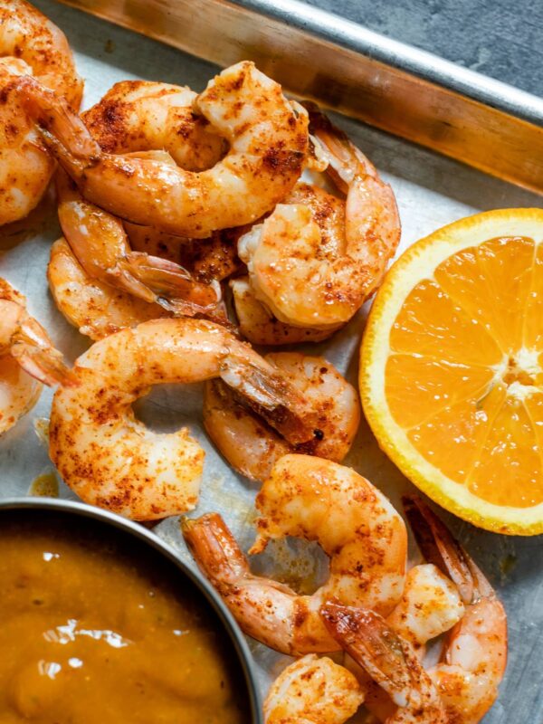 Pieces of chipotle shrimp on roasting sheet with slice of orange and bowl of mango sauce on a gray concrete surface
